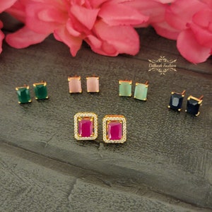 May include: A set of six pairs of gold stud earrings with different colored gemstones. The earrings feature a square-shaped setting with a clear crystal border. The gemstones are green, pink, light green, and blue.