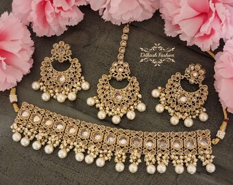 May include: A gold-tone Indian bridal jewelry set with clear stones and pearl accents. The set includes a choker necklace, earrings, and a maang tikka.