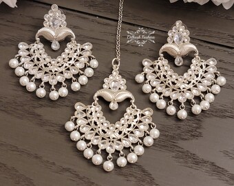 May include: A silver and white bridal jewelry set with a maang tikka, earrings, and a necklace. The set features intricate designs with clear stones and pearl accents.