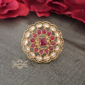 May include: A gold-toned ring with a central square ruby surrounded by teardrop-shaped rubies and clear, oval-cut stones. The ring has a detailed, circular design and is set against a gray background. The ring is a piece of jewelry.