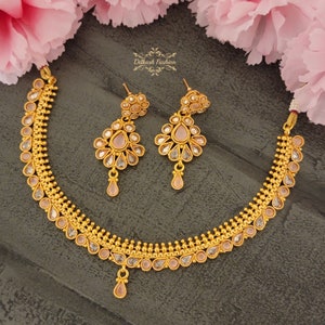 May include: A gold-toned necklace and earring set with pink and clear gemstones. The necklace and earrings have a floral design. The image includes the text "Dilkash Fashion". The jewelry is displayed on a dark gray surface with pink flowers in the background.