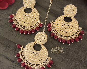 May include: Gold-toned jewelry set featuring a pair of large, round earrings and a headpiece. The earrings and headpiece are adorned with small white stones and dangling red beads. The set has a traditional, ornate design.
