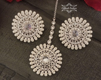 May include: A set of three silver-toned, round, jeweled ornaments with white pearl accents. The ornaments are arranged on a dark wood surface. The text "Dilkash Fashion" is visible in the top right corner of the image.
