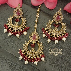 May include: A gold-tone Indian bridal jewelry set with red and white beads. The set includes a maang tikka, a necklace, and earrings.