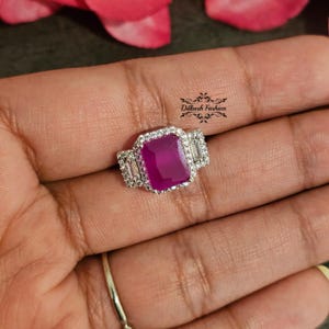 Peut inclure: Une bague en argent avec une grande pierre précieuse rose carrée entourée de petites pierres précieuses claires. La bague est sur le doigt d'une personne.