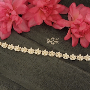 May include: A gold chain with white stones in the shape of flowers. The chain is on a dark gray surface with pink flowers in the background.