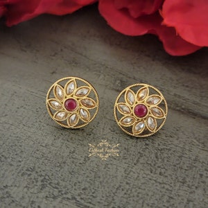 May include: A pair of gold-tone stud earrings with a floral design. Each earring features a circular base with a central red gemstone surrounded by smaller clear gemstones.