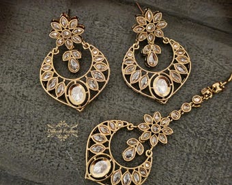 May include: A set of gold-tone earrings and a matching maang tikka, all adorned with clear sparkling stones. The earrings feature a floral design at the top and a crescent moon shape with intricate detailing. The maang tikka has a similar design with a floral top and a crescent moon shape.