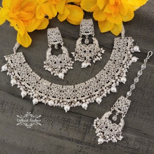 May include: A silver necklace set with a choker, earrings, and a maang tikka. The set features intricate floral designs with clear stones and pearl accents.