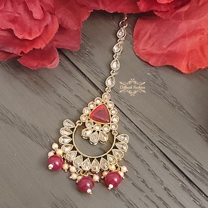 May include: A gold-tone maang tikka with a red gemstone and clear rhinestones. The maang tikka has a crescent moon shape with dangling red beads.