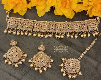 May include: A gold-tone Indian bridal jewelry set with a choker necklace, earrings, and a maang tikka. The set is adorned with clear stones and pearls.