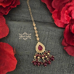 May include: A gold-toned headpiece with a teardrop-shaped, ruby-colored centerpiece and dangling ruby beads. The headpiece is adorned with small, round gold accents and a delicate chain. The background features red fabric.
