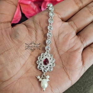 May include: A silver-toned headpiece with clear and red gemstones and a dangling pearl. The headpiece has a floral design with a central red teardrop-shaped stone. The brand name "Dilkash Fashion" is printed on the image.