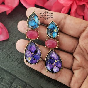 May include: A pair of long, dangling earrings with a gold-colored frame. Each earring features three sections: a teardrop-shaped blue stone, a pink oval stone, and a teardrop-shaped purple stone. The earrings are held in a hand.