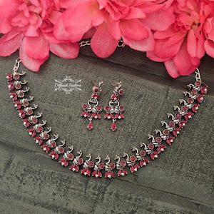 May include: A silver and red necklace and earrings set featuring a peacock design. The necklace has a row of red stones and a row of silver peacocks. The earrings are dangle earrings with a red stone and a silver peacock.