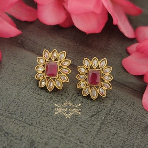 May include: A pair of gold-tone stud earrings with a square-shaped red gemstone surrounded by smaller clear gemstones. The earrings are set against a brown background.