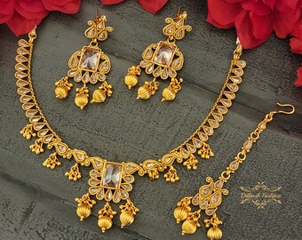 May include: A gold-tone necklace set with a teardrop-shaped pendant, matching earrings, and a matching maang tikka. The set features clear stones and intricate detailing.