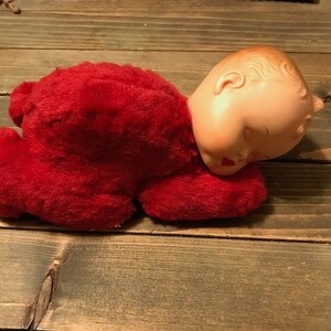 May include: A red plush baby doll with a plastic head, lying on its stomach with its eyes closed. The doll is wearing a red plush outfit.