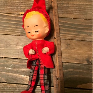 May include: A red and white plaid elf doll with a red hat and closed eyes. The doll is standing on a wooden surface.