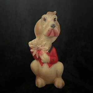 May include: A vintage, cream-colored, rubber dog figurine wearing a red jacket and holding a pink flower. The dog has a black nose and brown eyes.