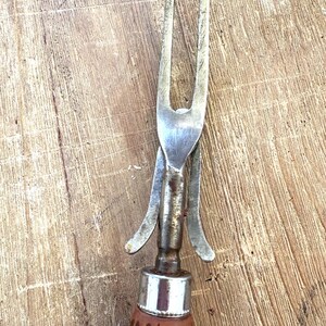 Puede incluir: Primer plano de un tenedor para trinchar vintage. Cuenta con una cabeza de acero inoxidable con dos p&uacute;as y un protector met&aacute;lico decorativo. El mango es de madera marr&oacute;n con un dise&ntilde;o acanalado y una tapa de metal plateado.