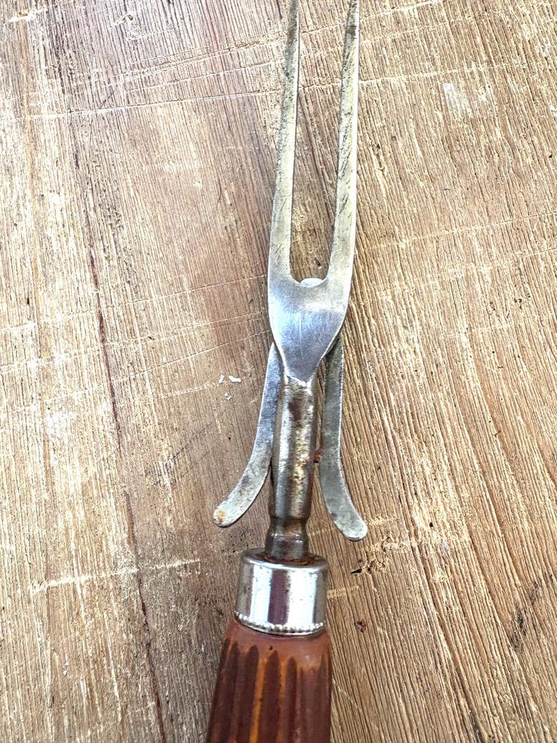 Puede incluir: Primer plano de un tenedor para trinchar vintage. Cuenta con una cabeza de acero inoxidable con dos p&uacute;as y un protector met&aacute;lico decorativo. El mango es de madera marr&oacute;n con un dise&ntilde;o acanalado y una tapa de metal plateado.