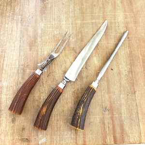 Puede incluir: Un juego de trinchar con tenedor, cuchillo y afilador. El juego presenta mangos de asta marrones con virolas de metal plateado. Las hojas del cuchillo y el afilador son plateadas. El tenedor tiene dos p&uacute;as.