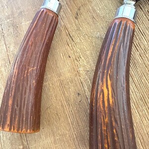 Puede incluir: Primer plano de dos cuchillos de trinchar antiguos. Los mangos son de color marr&oacute;n oscuro con un dise&ntilde;o texturizado y acanalado. Las virolas met&aacute;licas son plateadas y las hojas son visibles. Los cuchillos est&aacute;n sobre una superficie de madera.