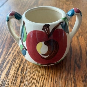 May include: A white ceramic creamer with two handles. The creamer is decorated with a red apple with a brown stem and green leaves. The apple has a yellow center.