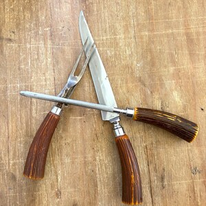 Puede incluir: Un juego de trinchar con cuchillo, tenedor y afilador. El cuchillo y el tenedor tienen hojas y p&uacute;as plateadas, con mangos texturizados marrones y naranjas. El afilador es plateado con un mango marr&oacute;n.