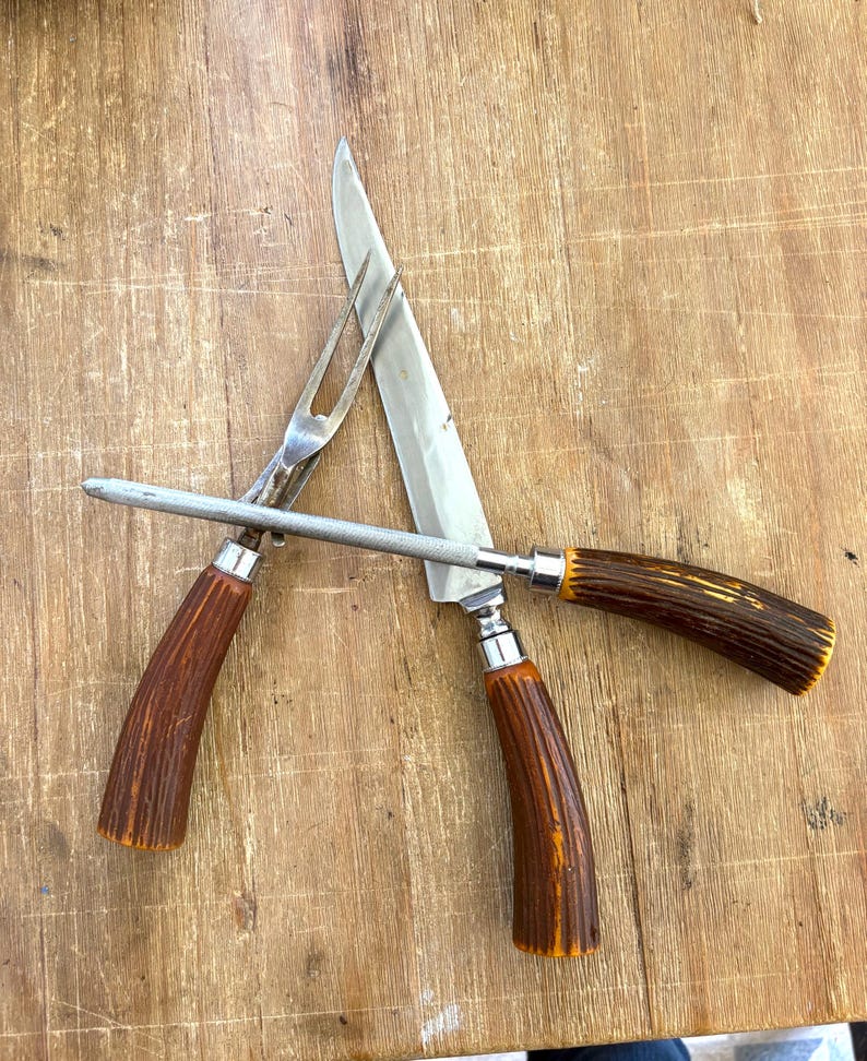 Puede incluir: Un juego de trinchar con cuchillo, tenedor y afilador. El cuchillo y el tenedor tienen hojas y p&uacute;as plateadas, con mangos texturizados marrones y naranjas. El afilador es plateado con un mango marr&oacute;n.