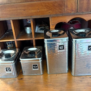 May include: Set of four stainless steel kitchen canisters with black lids. The canisters are labeled for coffee, tea, flour, and sugar. The canisters have a textured, hammered metal finish and are arranged in a row.