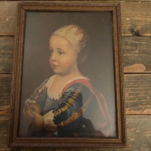 May include: A framed portrait of a young person wearing a white bonnet and a blue and red dress. The portrait is set against a dark background.