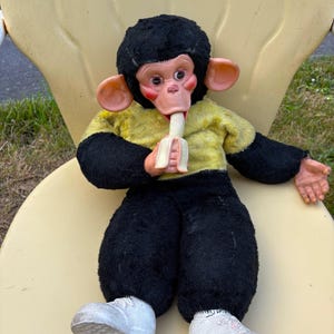 May include: A vintage monkey doll with a black fur head and body, wearing a yellow shirt, black pants, and white sneakers. The doll has a pink face and is holding a banana. The doll is sitting on a yellow chair.