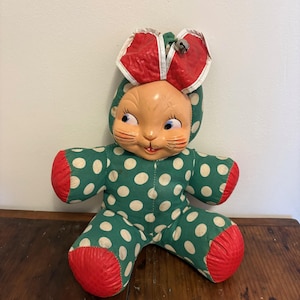 May include: Vintage stuffed bunny doll with a green and white polka dot jumpsuit and red accents. The bunny has a painted face, red ears, and a small bell on its head. A classic toy.