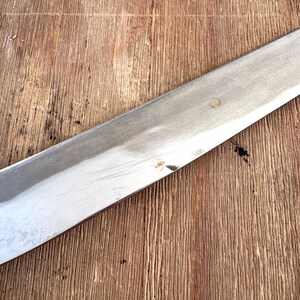 Puede incluir: Una hoja larga de acero inoxidable con un borde afilado, parcialmente visible. La hoja est&aacute; en &aacute;ngulo sobre una superficie de madera, contrastando el metal brillante con la veta texturizada de la madera. La madera tiene un tono marr&oacute;n c&aacute;lido.