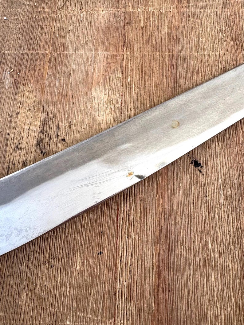 Puede incluir: Una hoja larga de acero inoxidable con un borde afilado, parcialmente visible. La hoja est&aacute; en &aacute;ngulo sobre una superficie de madera, contrastando el metal brillante con la veta texturizada de la madera. La madera tiene un tono marr&oacute;n c&aacute;lido.
