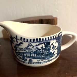 May include: A white ceramic creamer with a blue and white train design. The creamer has a handle and a spout. The train is a steam engine with a long line of cars.