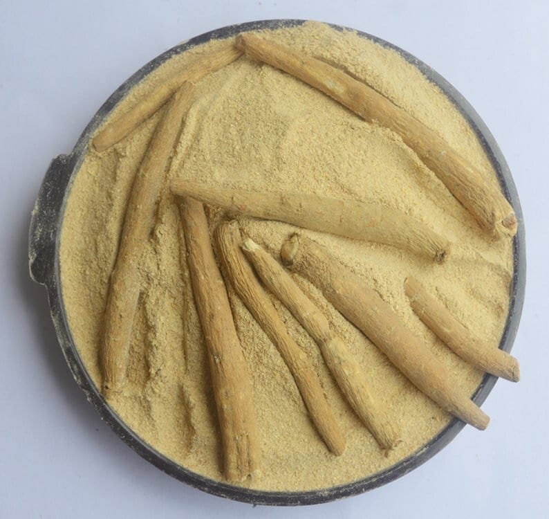 May include: A close-up of a black bowl filled with light brown powder and several long, thin, light brown sticks. The sticks are arranged in a radial pattern, with the tips pointing towards the center of the bowl.