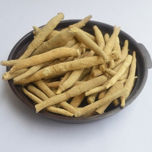 May include: A close-up of a bowl filled with dried, light brown, thin, and long herbal roots. The roots are arranged in a random pattern and are piled high in the bowl.