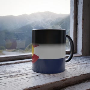 May include: A black color-changing mug with a design of the Colorado state flag. The mug is sitting on a wooden surface with a window and a mountain view in the background.