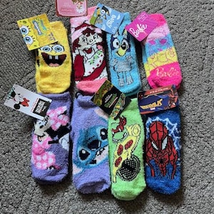 May include: A selection of colourful, fluffy socks featuring cartoon characters such as SpongeBob, Bluey, and Spider-Man. Other socks display designs of Strawberry Shortcake, Barbie, Stitch, and a pizza. Each pair has a tag.