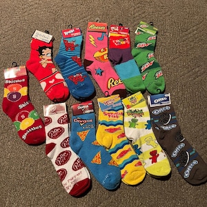 May include: A selection of patterned socks featuring popular brand logos. Designs include Skittles, Betty Boop, Reese's, Doritos, Dr. Pepper, Swedish Fish, and Oreo. The socks are arranged on a plain, neutral-toned surface.