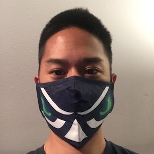 May include: Navy blue face mask with a white and green graphic design. The mask has a geometric pattern with a stylized eye shape. The mask is made of fabric and covers the nose and mouth.
