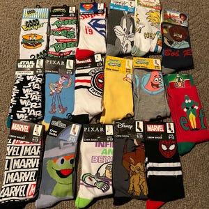 May include: A collection of crew socks featuring various cartoon characters and logos. Designs include characters from Looney Tunes, Marvel, Disney, Pixar, and Sesame Street. The socks are in various colors and sizes 9-13.