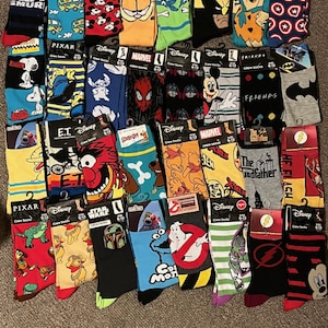 May include: A collection of colorful crew socks featuring various cartoon characters and pop culture icons. Designs include characters from Disney, Marvel, and other popular franchises. The socks are displayed on a neutral surface.