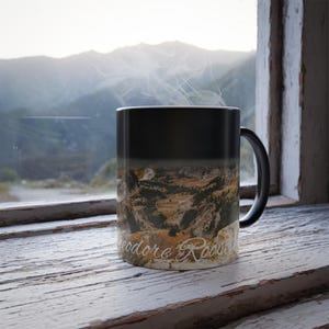 May include: A black magic mug with a scenic image of a mountain range. The mug is filled with a hot beverage and steam is rising from the top. The text "Theodore Roosevelt" is printed on the bottom of the mug.