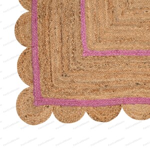 May include: A square jute rug with a pink border and a scalloped edge. The rug is made of natural fibers and has a rustic, earthy look.