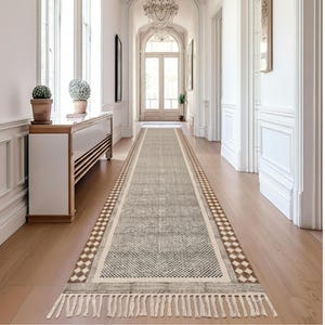 Puede incluir: Una alfombra de pasillo rectangular gris y beige con un patrón geométrico y flecos. La alfombra tiene un borde con un patrón de diamantes repetido en marrón y blanco. La alfombra está colocada sobre un suelo de madera en un pasillo.