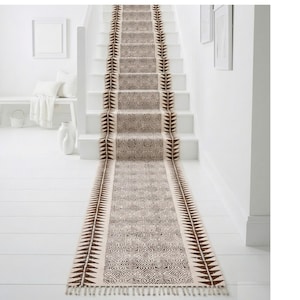 Puede incluir: Una alfombra de escalera con estampado beige y marrón con un diseño geométrico. La alfombra presenta un patrón repetitivo de triángulos marrones a lo largo de los bordes y un diseño central detallado. La alfombra está colocada en escaleras blancas.
